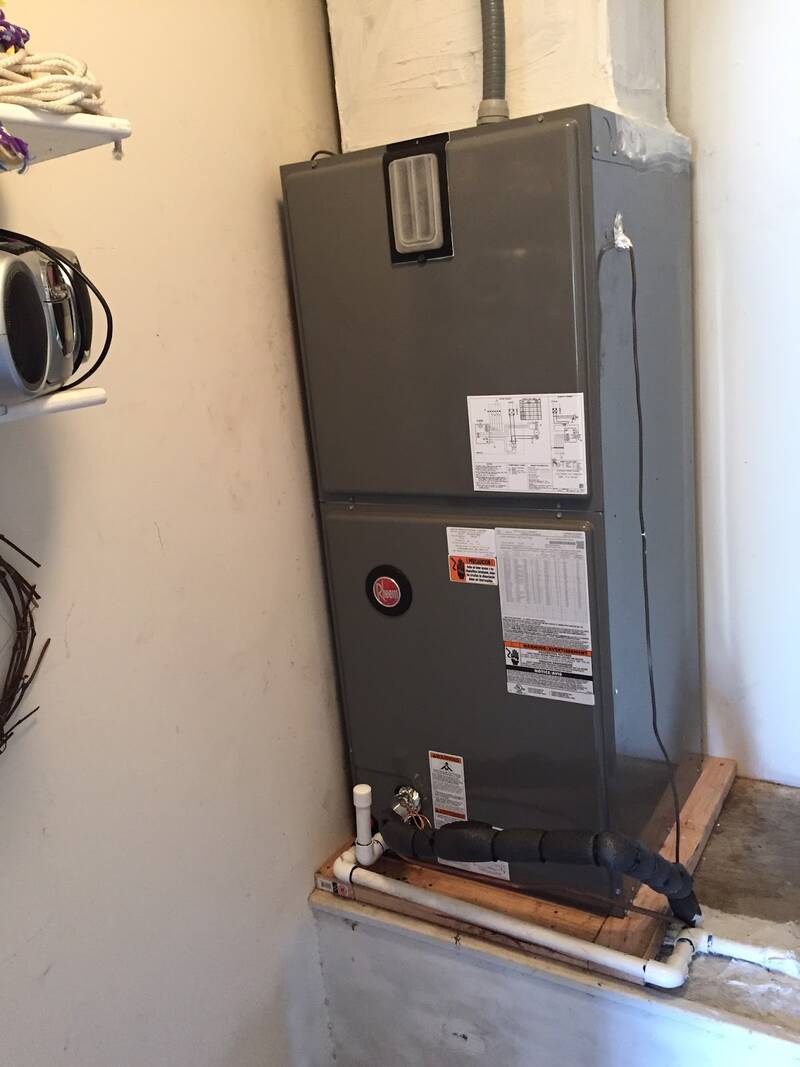 Grey Rheem air handler on a wood platform in a utility closet, with copper line set and white drain pipe