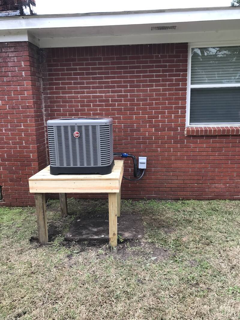 Rheem condenser on a new raised wooden stand beside a red brick house, under a window
