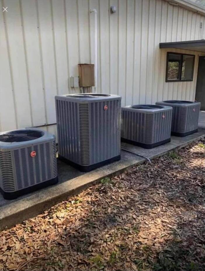 Four grey Rheem condensers in a row on a concrete slab beside a metal-sided building