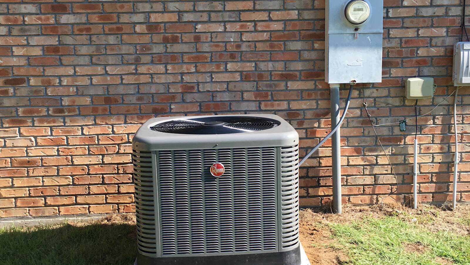 New grey Rheem condenser on a pad against a red brick wall, next to the electric meter